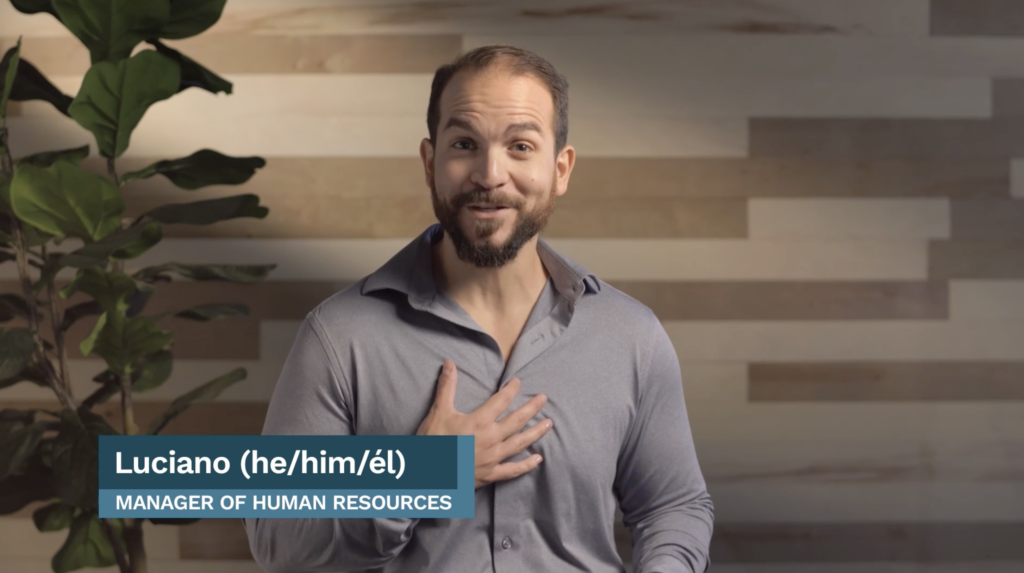Luciano (he/him/él), Manager of Human Resources, smiles and gestures to himself in front of a wood-patterned wall